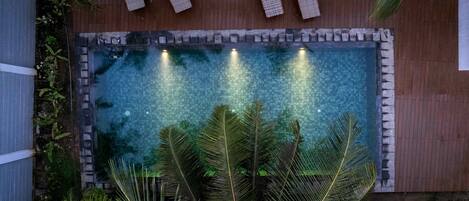 Una piscina rectangular con borde de azulejos, rodeada por una terraza de madera y palmeras.