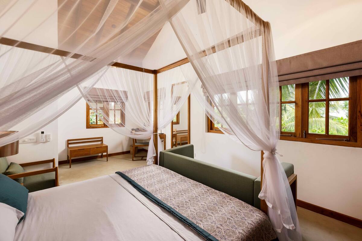 Un dormitorio con cama, banco, mesita de noche y ventana con vistas a la vegetación.