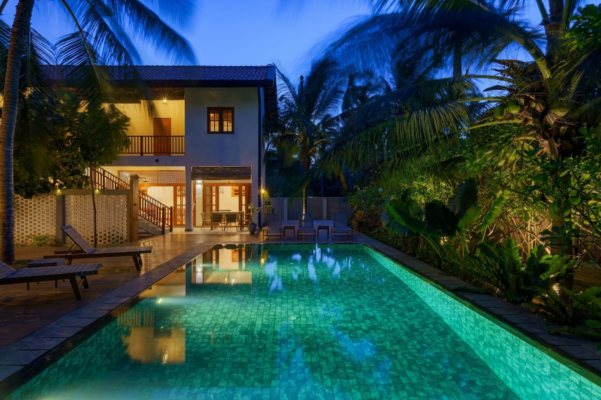 Una casa de dos pisos con piscina, rodeada de palmeras y abundante vegetación.