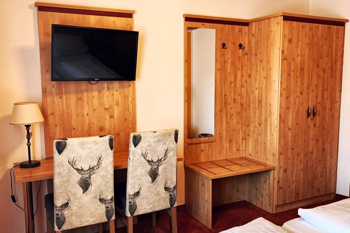 A hotel room with a wooden desk, two chairs with deer head designs, a television, a wardrobe, and a bed.