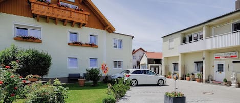 Un hôtel de style architectural européen traditionnel, doté d’un balcon en bois orné de jardinières, d’une allée pavée et d’une voiture garée devant.