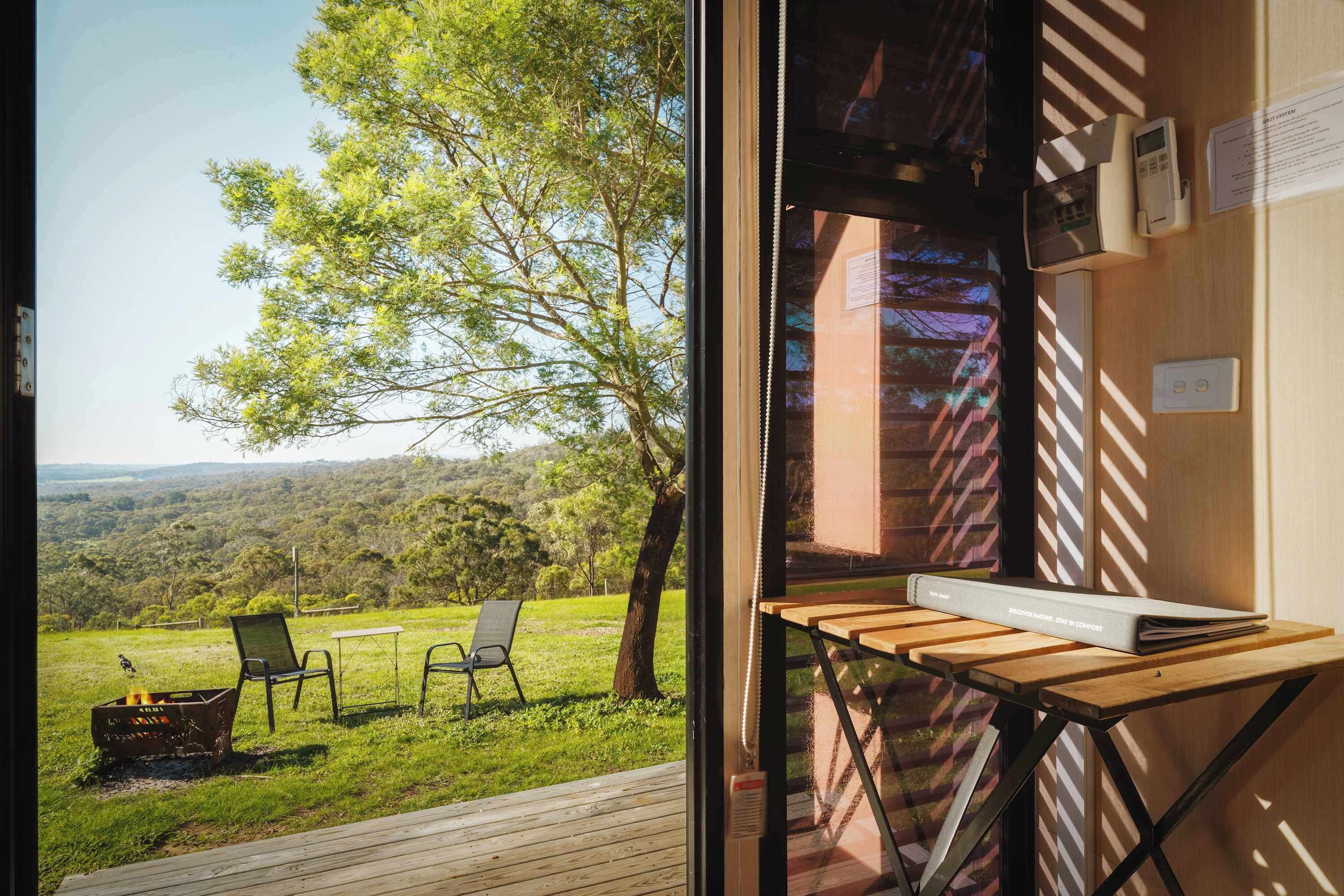 A room with a view of a grassy landscape and trees. There is a table with a book and a chair.