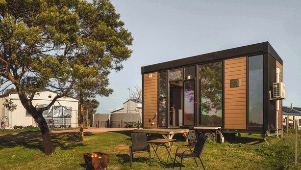 A modern, single-story cabin with large windows, a patio with a fire pit, and a tree in the foreground.