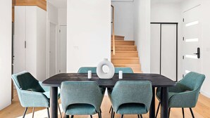 Ein moderner Essraum mit einem dunklen Holztisch, türkisfarbenen Stühlen, einer weißen Vase und einer Treppe im Hintergrund.