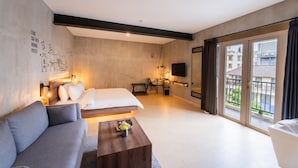 A modern hotel room with a bed, a sofa, a coffee table, a TV, and a balcony with a view of buildings. - The All New Stark Boutique Hotel (Kuta)
