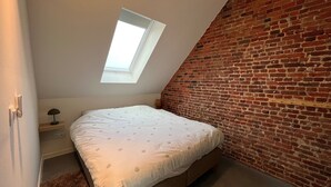 Una camera da letto con soffitto a falde, un lucernario, una parete in mattoni, un letto con materasso bianco e un comodino con una lampada.