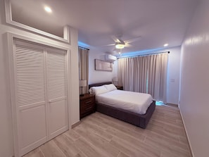Ein Schlafzimmer mit einem Bett, einem Deckenventilator, einem Nachttisch, einem Kleiderschrank und einem Fenster mit Vorhängen.
