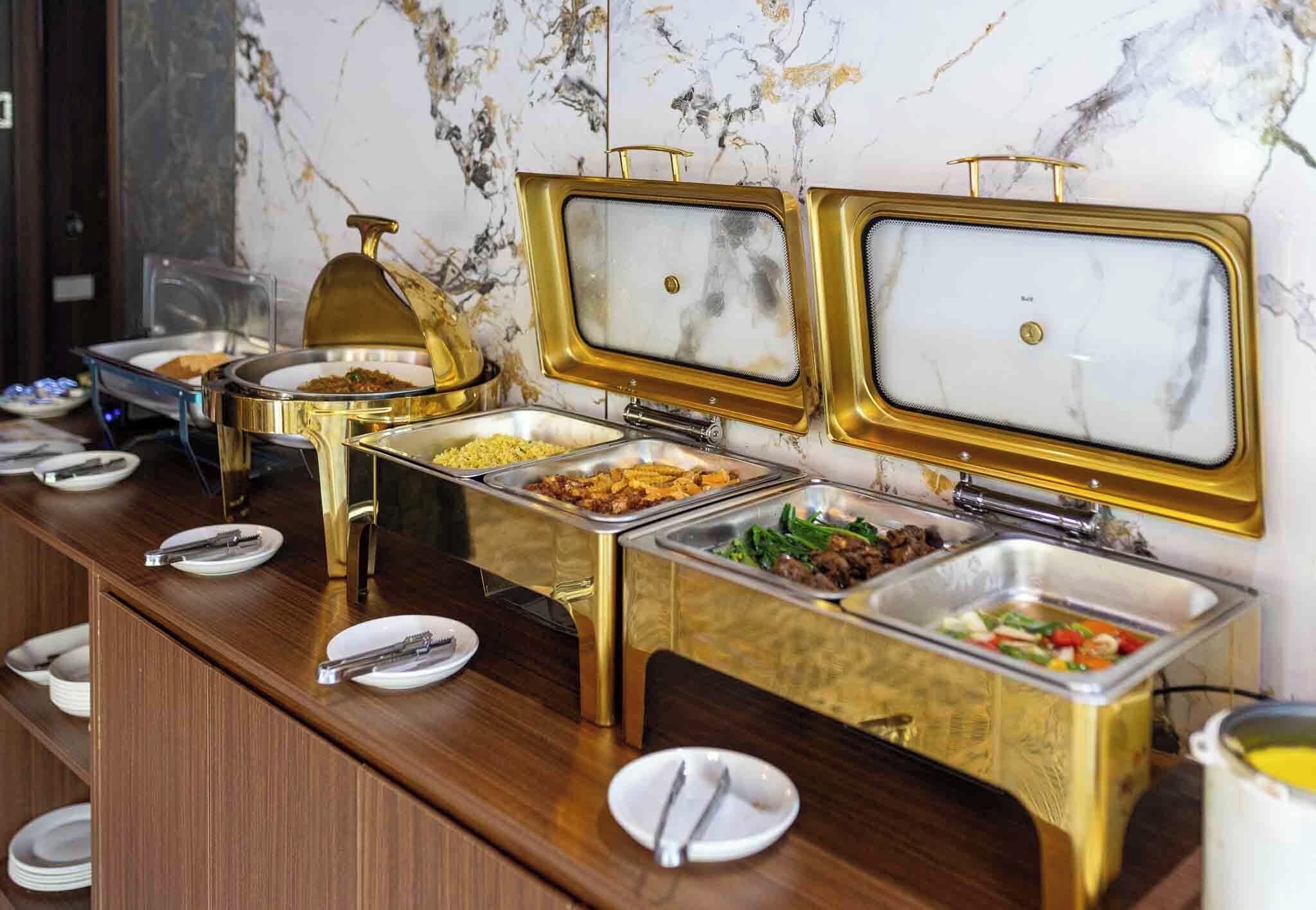 A buffet with multiple food stations, each with a gold-colored serving tray and a glass cover.