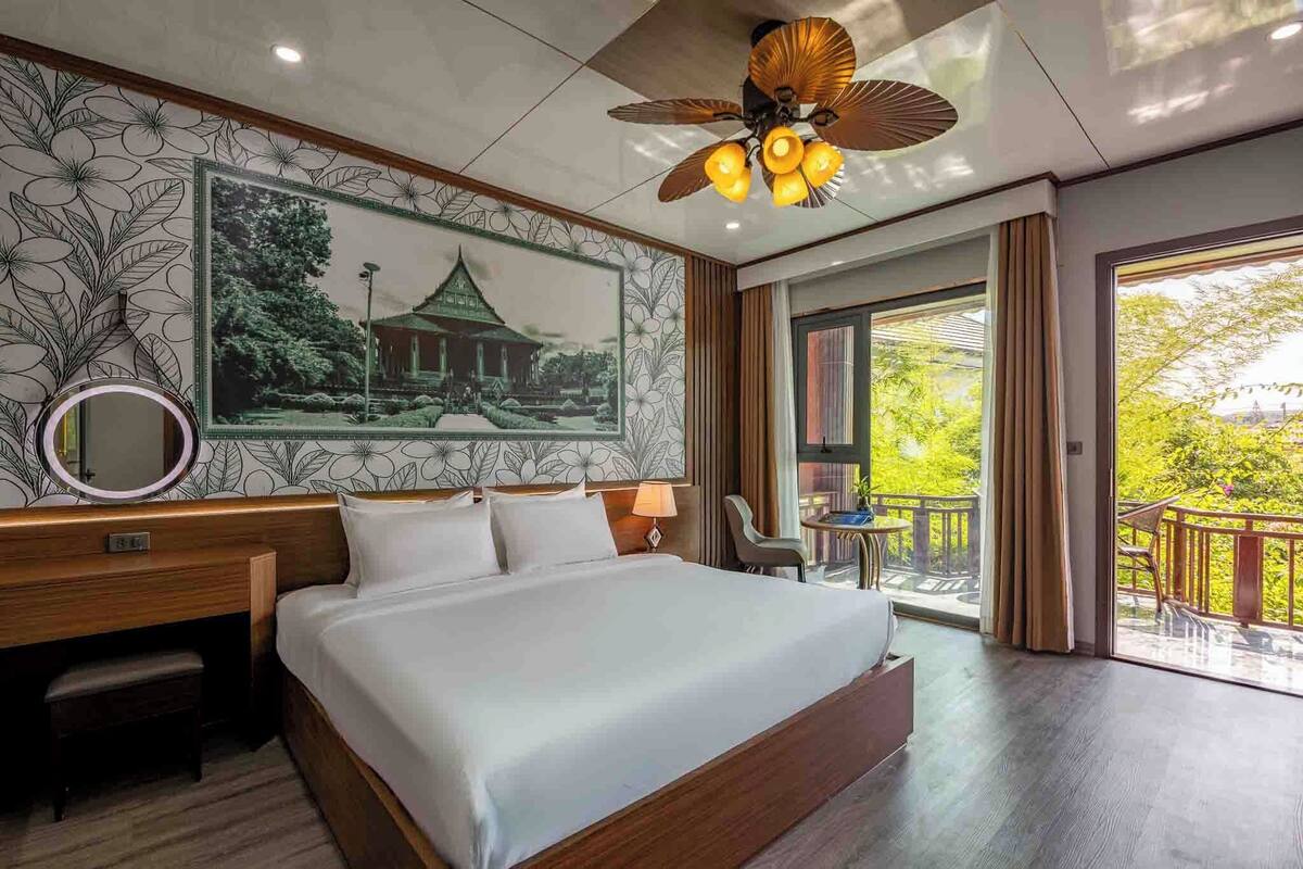 Habitación de hotel con una cama grande, un escritorio, una silla, un balcón con vistas a la vegetación y un ventilador de techo.