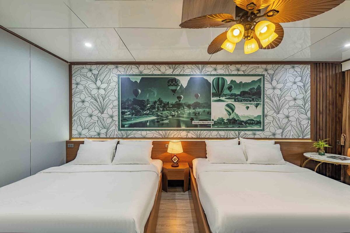Habitación de hotel con dos camas, ventilador de techo, lámpara de pared y un cuadro grande con globos aerostáticos.