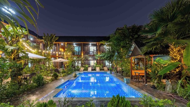 A pool area with lounge chairs and umbrellas, surrounded by lush greenery and a building with multiple windows and outdoor seating. - Champa Phan Boutique Hotel (Vientiane)