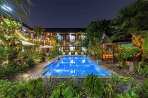 A pool area with lounge chairs and umbrellas, surrounded by lush greenery and a building with multiple windows and outdoor seating. - Champa Phan Boutique Hotel (Vientiane)