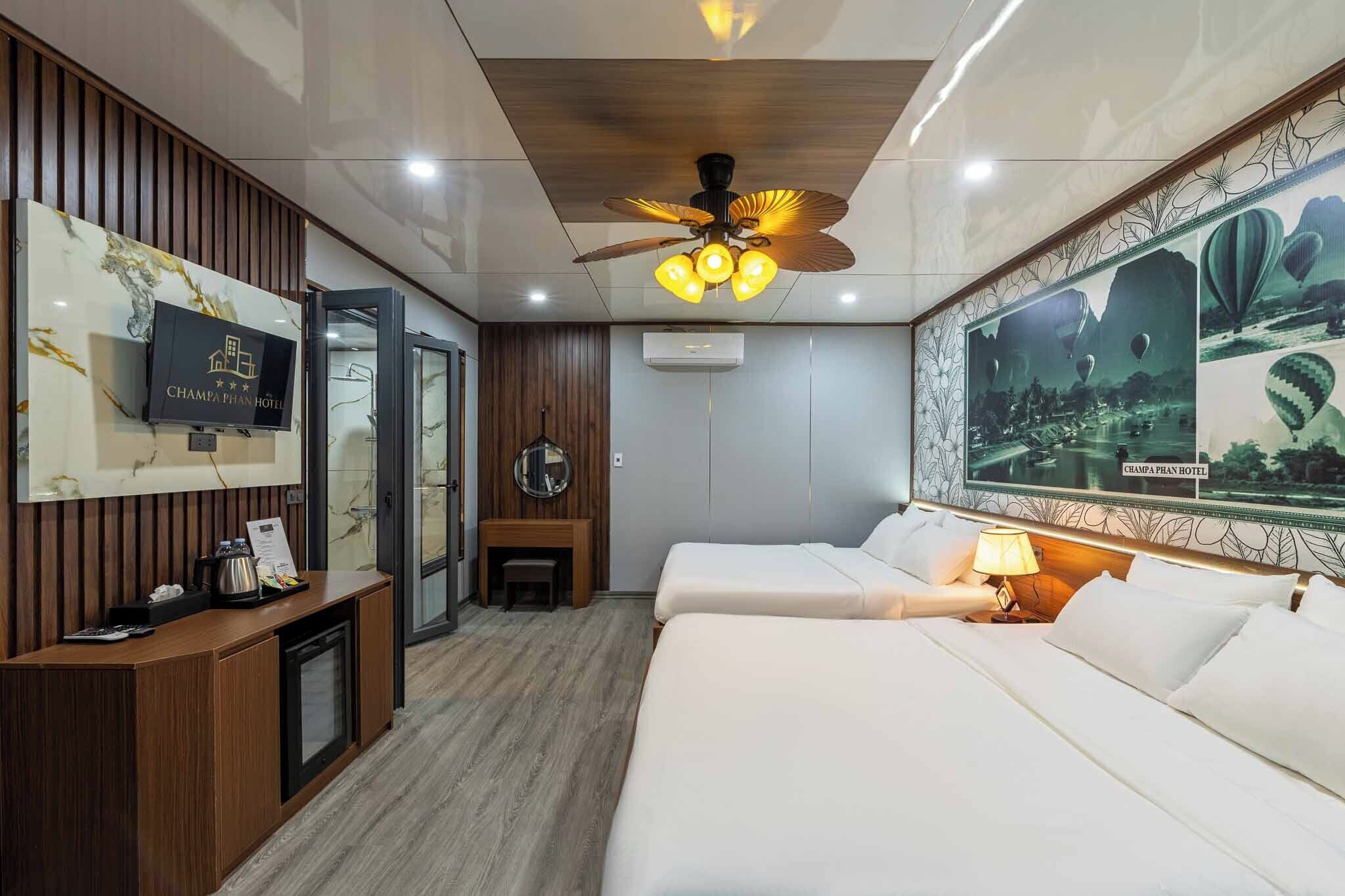 A hotel room with a wooden reception desk, a television, a ceiling fan, a bed, a bedside table with a lamp, and a large framed picture of hot air balloons.