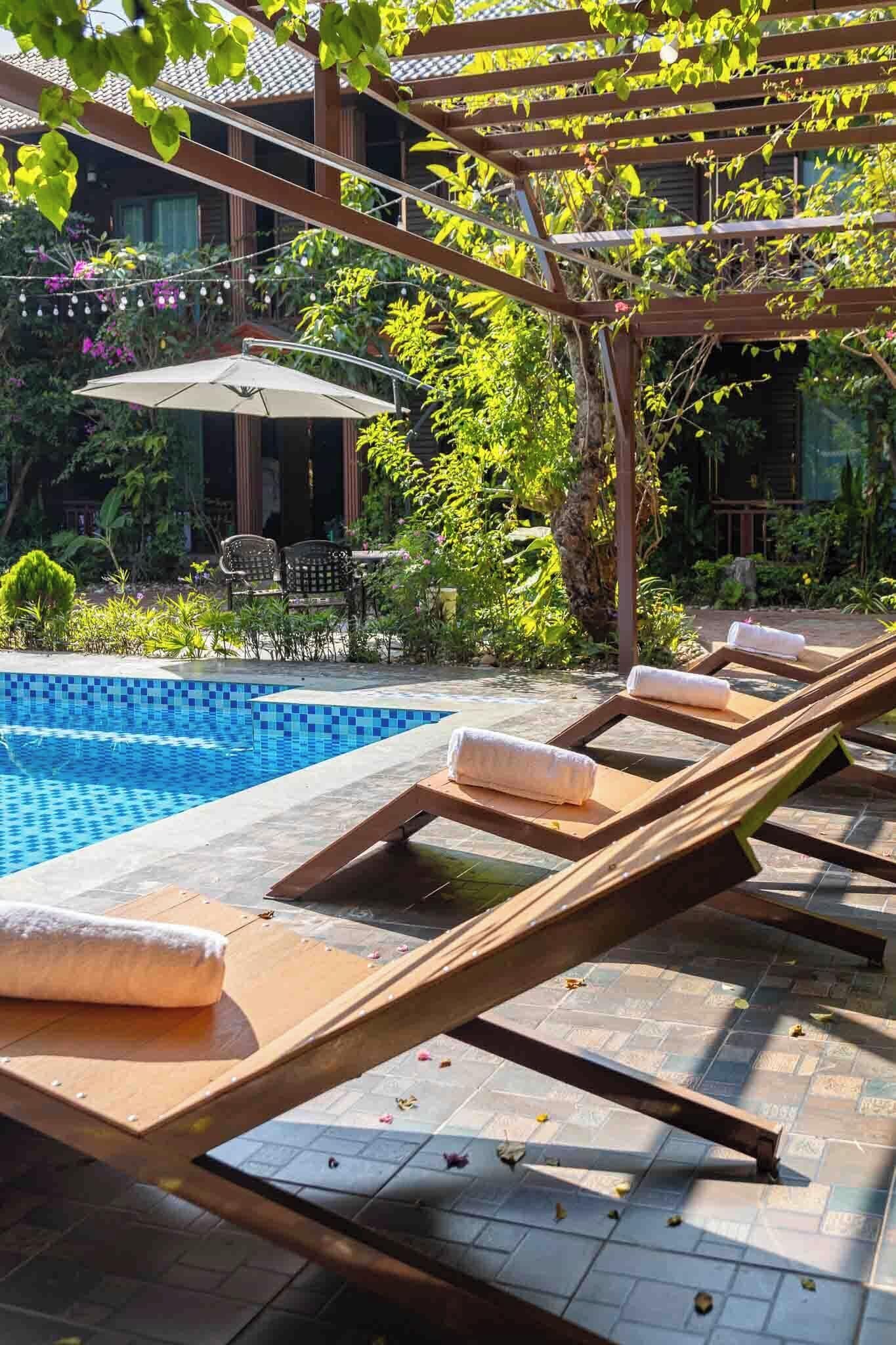 Een zwembadterras met ligbedden, een parasol en een pergola begroeid met planten.