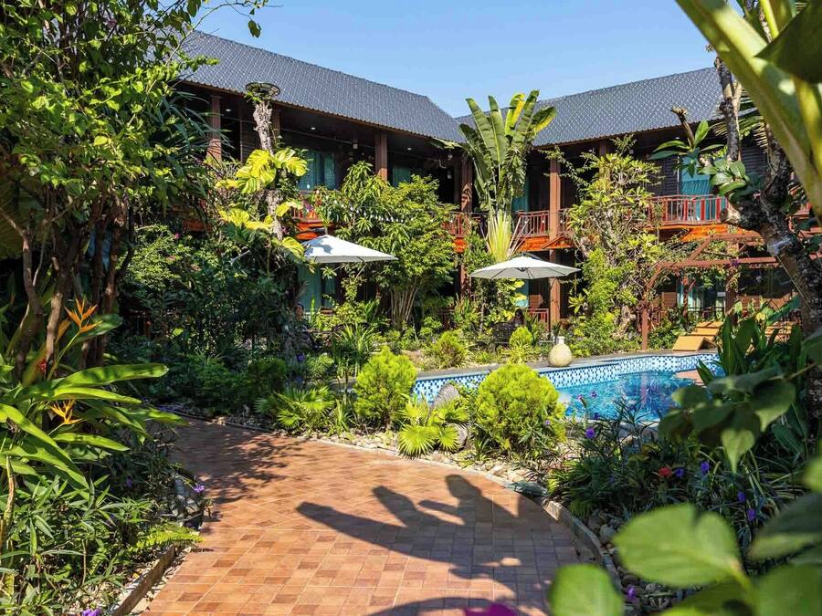A lush garden with a pool, wooden buildings with balconies, and a brick pathway.