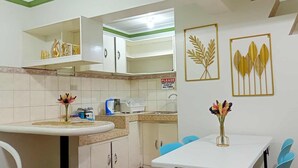 Une cuisine compacte avec des meubles blancs, un évier et un coin repas agrémenté de chaises bleues. On y trouve également des éléments décoratifs muraux et un panneau rappelant de garder la cuisine propre.