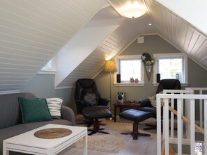 Ein gemütliches Dachzimmer mit schrägen Wänden, einem grauen Sofa, einem schwarzen Ledersessel, einem weißen Tisch und einem weißen Geländer.