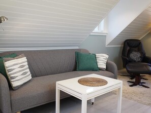 En grå soffa med gröna och randiga kuddar, ett vitt bord med en vävd bordsduk och en svart läderfåtölj med en kudde med ett lövmönster.