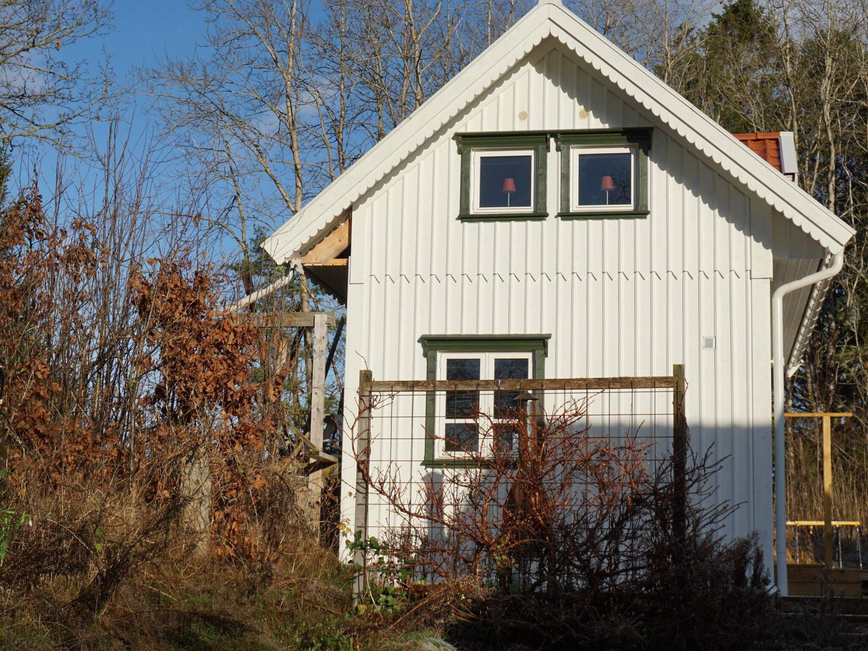 A white building with a gabled roof, two windows with shutters, and a wooden fence in front.