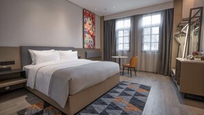 Ein modernes Hotelzimmer mit einem großen Bett, einem Schreibtisch und einem Sessel. Ein Fenster mit Vorhängen, ein Bild an der Wand und ein Spiegel auf dem Nachttisch.