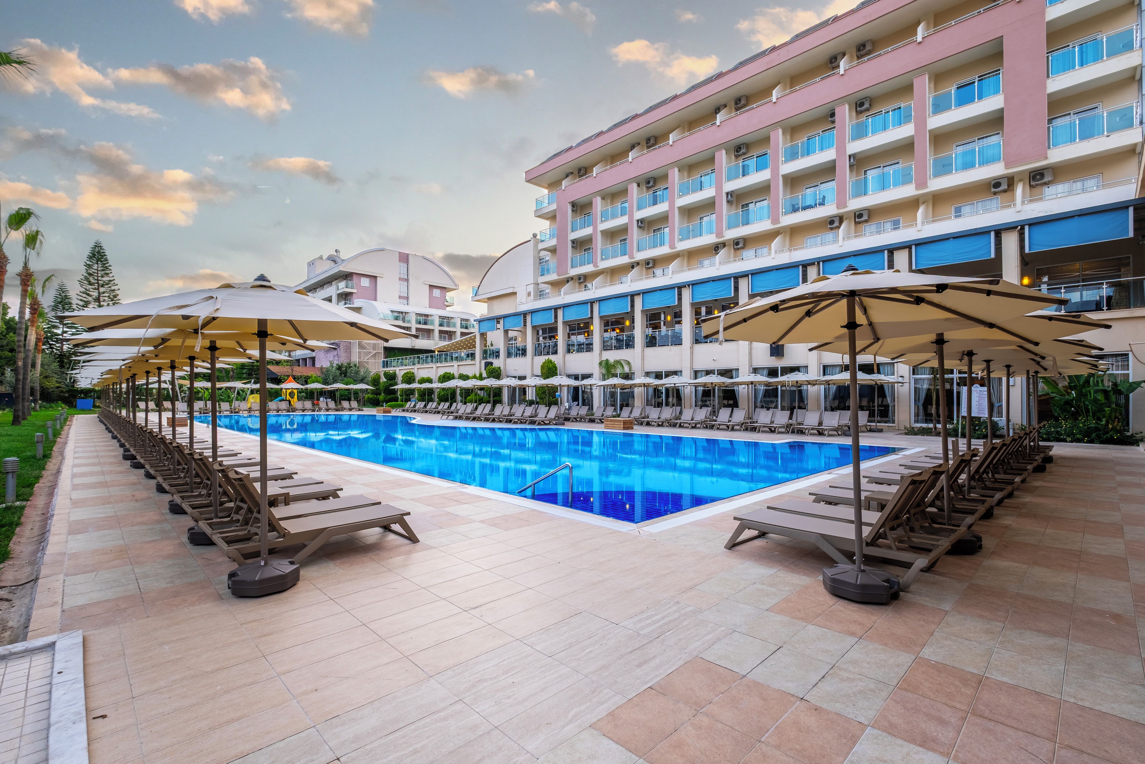 Zona de piscina hotelera con tumbonas y sombrillas, rodeada de un edificio con varios balcones.