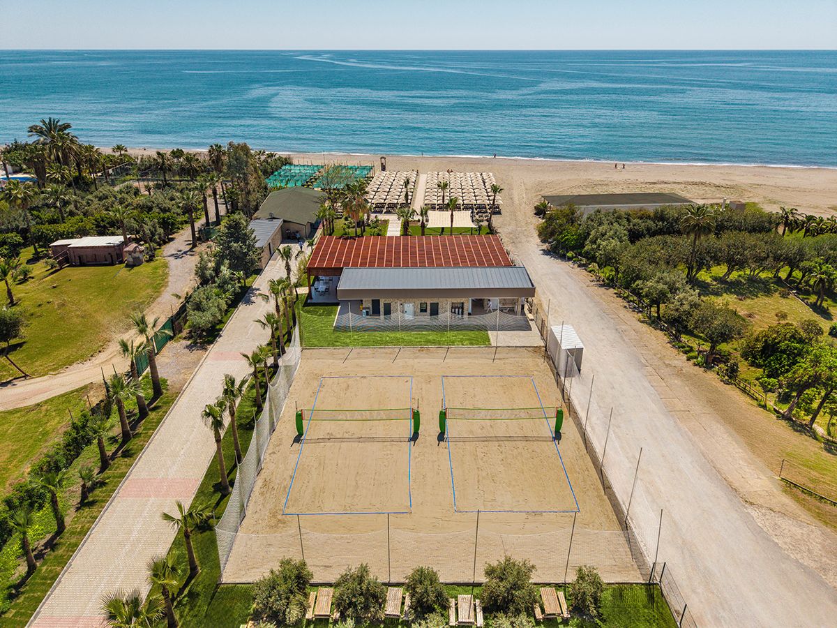Una propiedad costera con cancha de tenis, un edificio con techo rojo y una playa al fondo.
