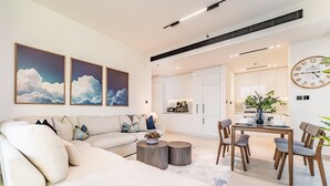 Een moderne woonkamer met een bank, salontafel en een eethoek met stoelen en een tafel. Er hangen drie schilderijen aan de muur en staat een klok.