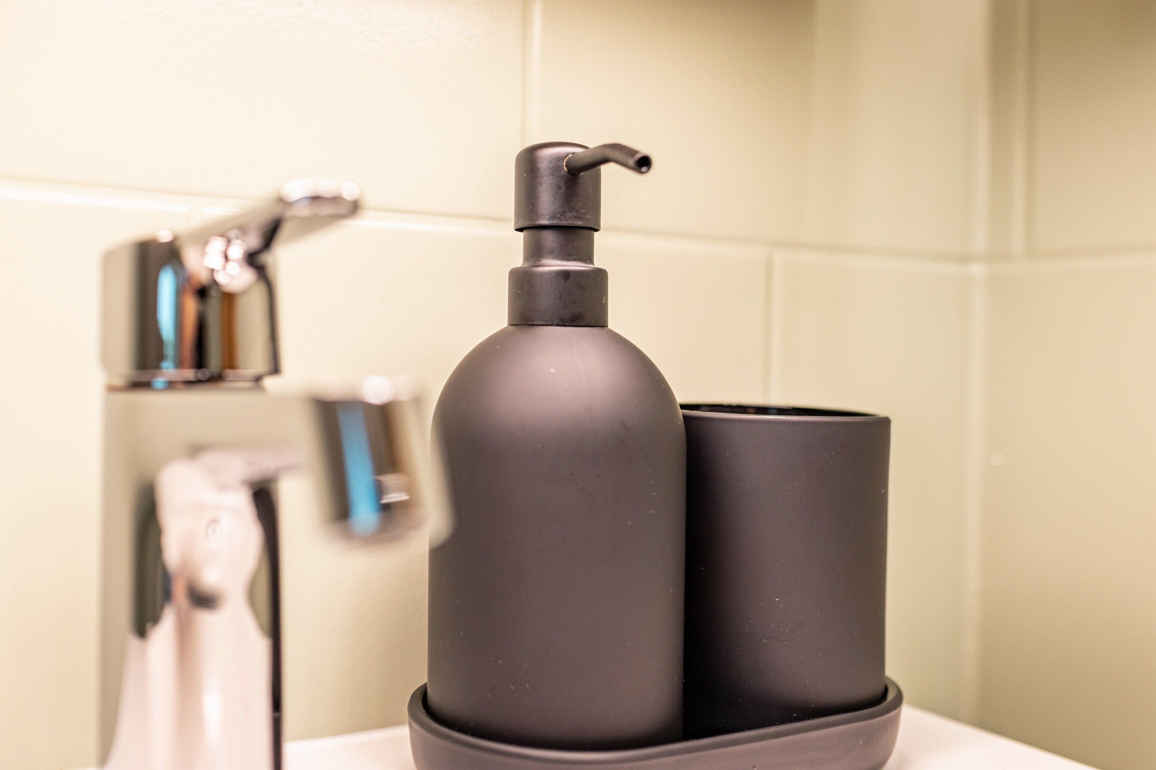 Un baño con una botella dispensadora negra y una taza en el lavamanos.