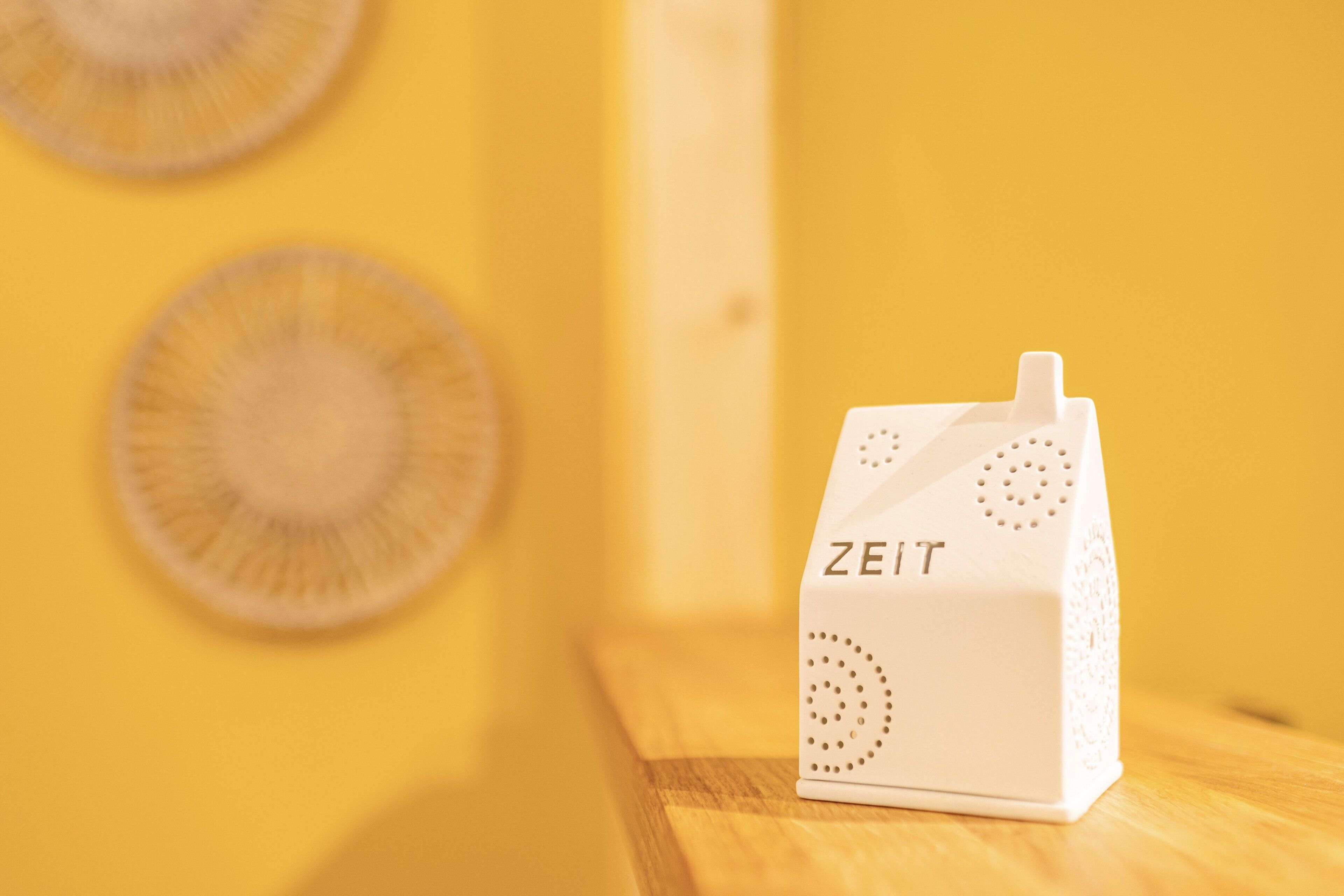 Een wit object met de tekst 'ZEIT' erop, geplaatst op een houten ondergrond voor een gele achtergrond.