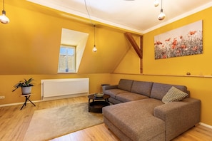 Een woonkamer met een hoekbank, een bijzettafeltje, een kamerplant, een radiator en een raam met uitzicht.