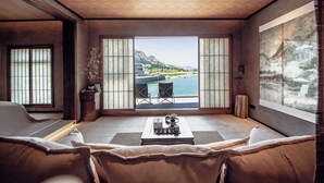 A room with a large sliding glass door leading to a balcony with a view of the sea and mountains. There is a sofa, a coffee table with a plant, and a small side table. - Nordsee Manor by Arcadia Collection (Qingdao)