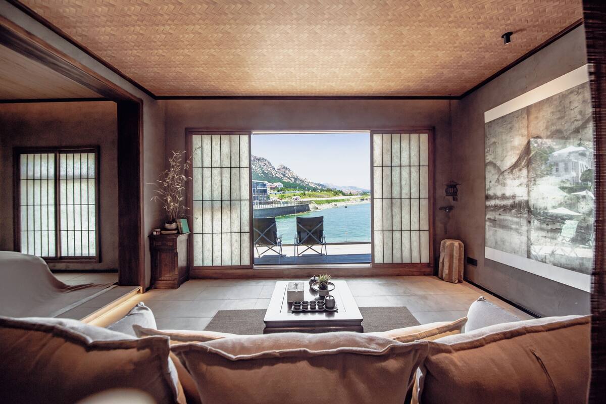 Habitación con un gran ventanal corredero que da a un balcón con vistas al mar y a las montañas. Se observa un sofá, una mesa de centro con una planta y una mesita auxiliar.