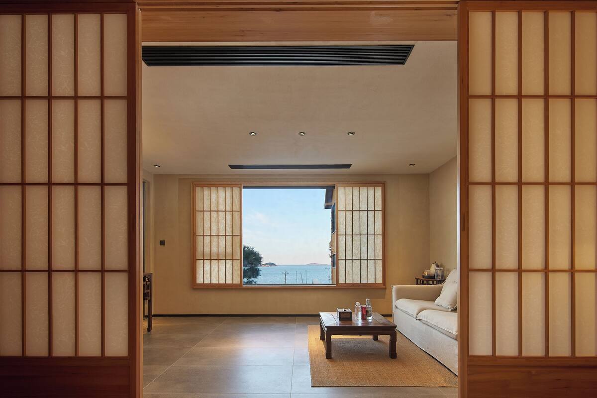 Una habitación tradicional japonesa con suelo de tatami, una mesa baja con cojín y vistas al mar a través de biombos shoji.