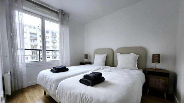Een hotelkamer met twee bedden, witte beddengoed, een raam met gordijnen, een nachtkastje met lamp en uitzicht op gebouwen.