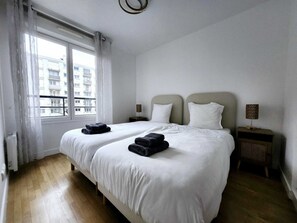 A hotel room with two beds, white bedding, a window with curtains, a bedside table with a lamp, and a view of buildings outside. - Exquisite Luxury Apartment in Charenton-le-Pont   (Charenton-le-Pont)