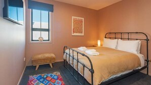 Une chambre avec un lit simple, une fenêtre, une œuvre d’art accrochée au mur, un banc et un tapis coloré.