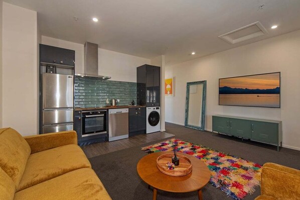 A modern kitchen with stainless steel appliances, a dining area with a wooden table, and a colorful rug. - Bridge St. Apartment 1 (Nelson)