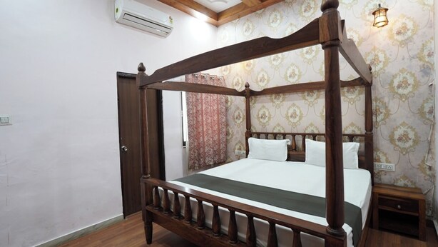 A four-poster bed with a wooden frame and a green bedspread, a nightstand with a lamp, and patterned wallpaper. - Nimbal Elegant Home (Jaipur)