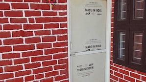 A red brick wall with a white door labeled "MADE IN INDIA" and a window with a mesh screen. - Nimbal Elegant Home (Jaipur)