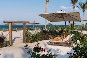 A beachside lounge area with a wooden pergola, a thatched umbrella, and a reclining chair.