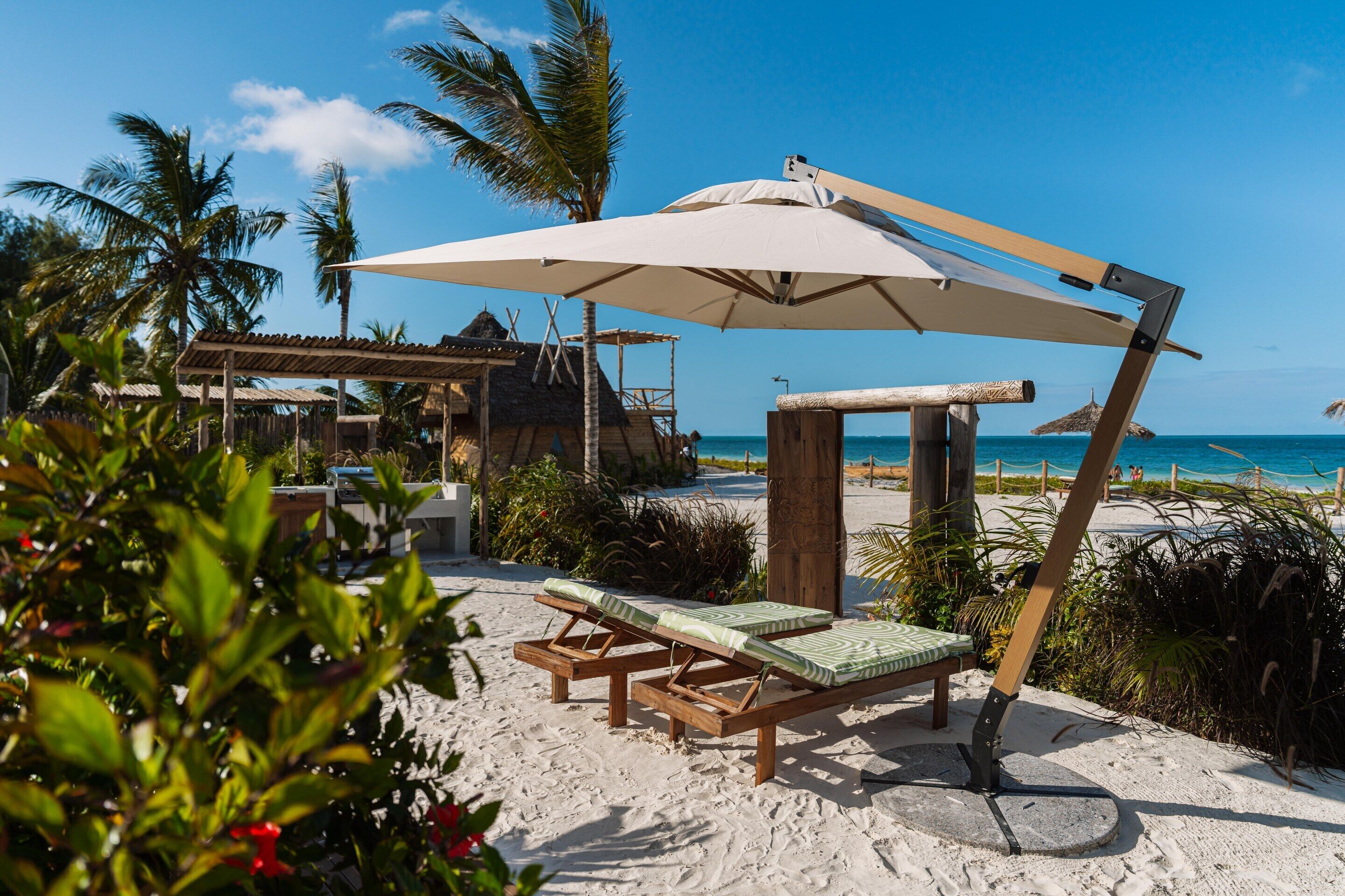 A beachside lounge with wooden lounge chairs and a large umbrella, surrounded by palm trees and a clear view of the ocean.