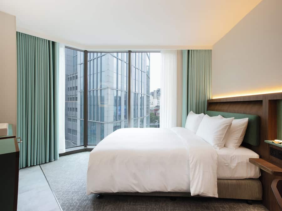 A modern hotel room with a large bed, a desk, and a view of a cityscape through floor-to-ceiling windows.