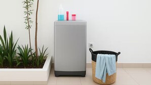 Un refrigerador gris con botellas de colores encima, un cesto de basura negro con un paño azul, y plantas verdes en una maceta blanca.