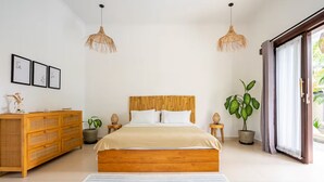 Un dormitorio moderno con una cama de madera, dos lámparas colgantes de ratán, un aparador de madera y un ventanal con vistas a la vegetación.