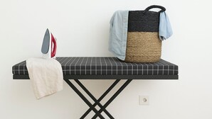 Una tabla de planchar con un planchador y un paño, una cesta tejida con un paño azul, y un enchufe en la pared.