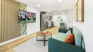 Una sala de estar moderna con un sofá verde, una mesa de centro y un televisor de pantalla plana mostrando un partido deportivo. Se ve una cocina al fondo.