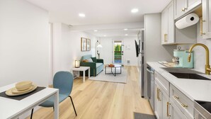 Una cocina moderna con armarios blancos, un lavamanos y una encimera. Hay una zona de comedor con una mesa y sillas, y un sofá al fondo.