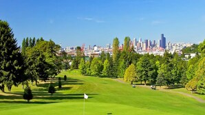 Campo de golf con una bandera, árboles y un horizonte urbano al fondo.