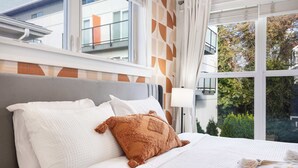 Ett hotellrum med ett stort fönster, en säng med vita sängkläder och utsikt över byggnader och träd.