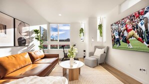 Ein modernes Wohnzimmer mit einem Ledersofa, einem runden Couchtisch, einem grauen Sessel und einem großen Fernseher, der ein Fußballspiel zeigt.